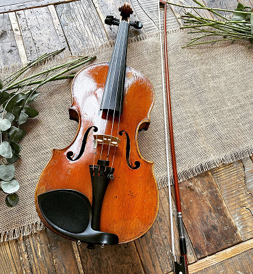 Brown violin and bow placed on rustic wood floor with burlap fabric and green foliage nearby.