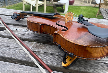 Violin and bow resting on weathered wooden picnic table outdoors with grass and chairs in background.
