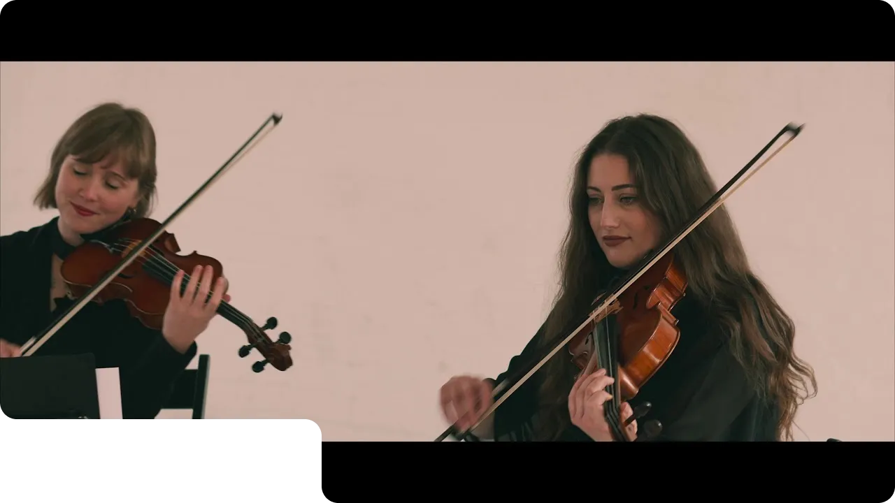 Two women playing violins indoors with focused expressions and bows in motion.