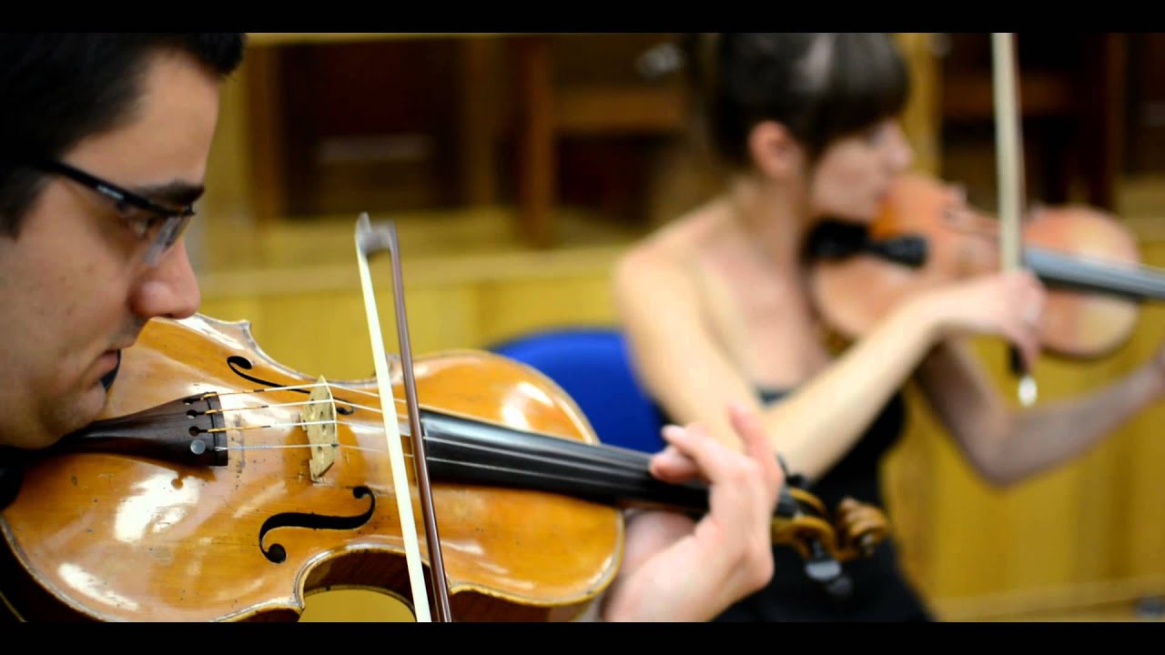 Close-up of a man playing violin with a woman playing violin blurred in the background.