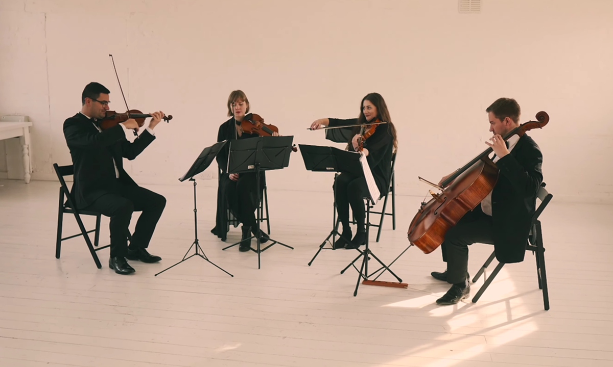A string quartet of four musicians seated in a minimal white room playing violins, viola, and cello.