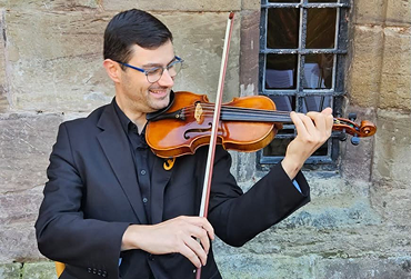 Smiling man in black suit playing violin beside rustic stone wall with window.
