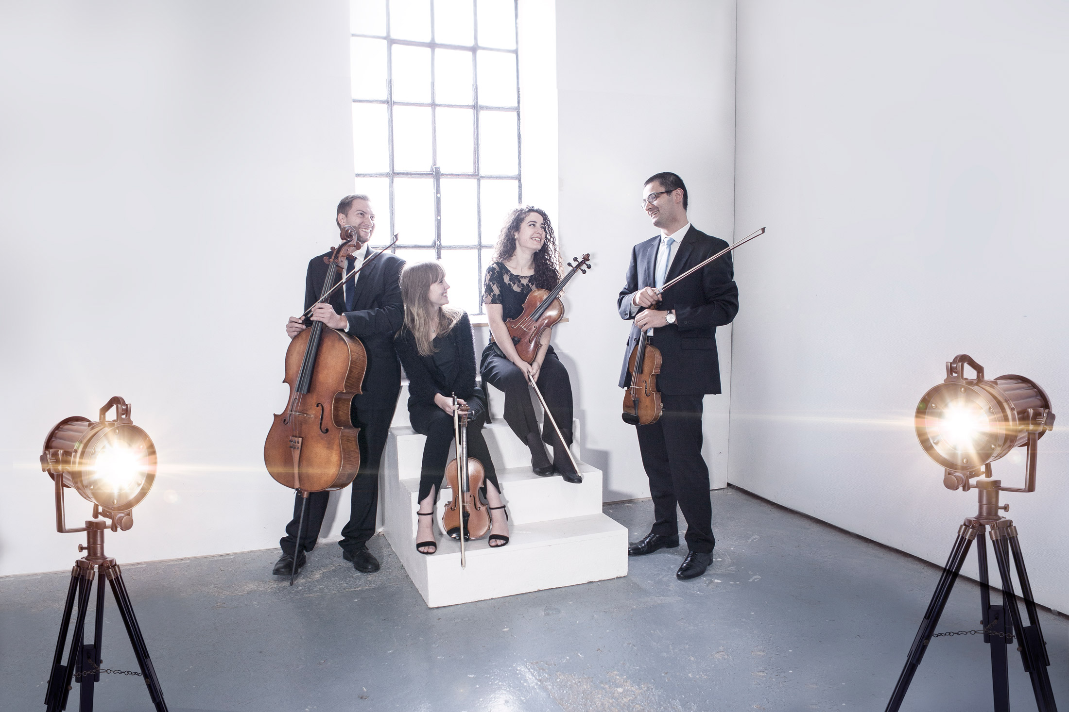 Four classical musicians holding string instruments in a bright studio with large window and spotlights.